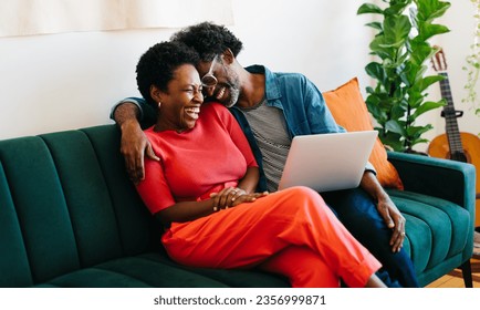 Una pareja feliz y madura, relajándose en casa, disfrutando de un tiempo de calidad en el sofá. Se ríen, abrazan y usan un portátil, creando un momento cálido y auténtico de amor y unión. Foto de stock