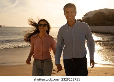 Happy mature couple running together on beach at sunset, cheerful middle aged man and woman holding hands and smiling, spending time on ocean shore, enjoying date or vacation trip, free space - Powered by Shutterstock