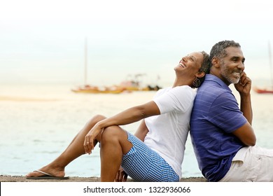 Happy Mature Couple Sitting Back To Back By The Beach.