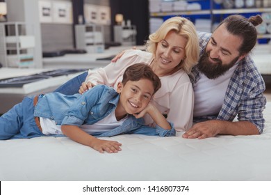 Happy Mature Couple Smiling At Their Cheerful Young Son, Lying Together On Orthopedic Mattress At Furniture Store. Lovely Family Enjoying Shopping For Furniture At Home Goods Shop