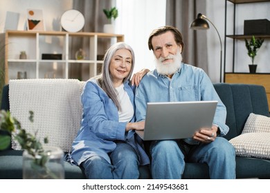 Happy Mature Couple Using Laptop Watching Movie Together Sitting On Couch At Home. Older Couple Browsing Internet On Computer Reading Online News On Weekend. Technology And Gadgets