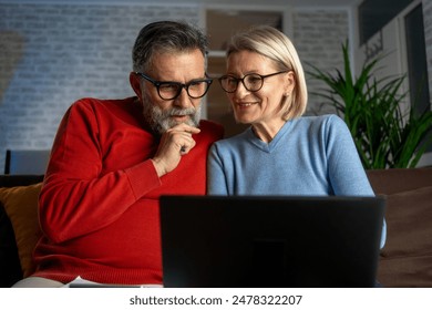 Happy mature couple using laptop at home. Happy couple with laptop and credit card sitting at home. Happy biracial couple sitting on sofa and using laptop in living room at home - Powered by Shutterstock