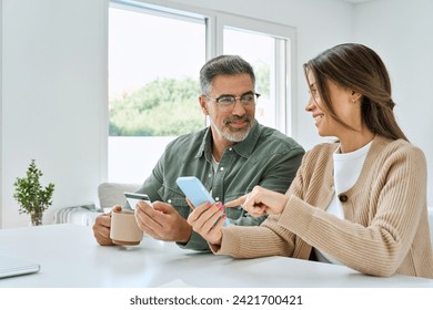 Happy mature couple using phone holding credit card making payments. Middle aged older man and woman doing ecommerce shopping on smartphone booking or buying online on mobile sitting at home table. - Powered by Shutterstock