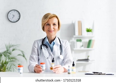 happy and mature doctor in white coat holding bottles with cbd and medical cannabis lettering