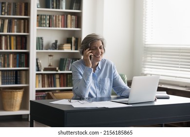 Happy Mature Elderly 50s Business Professional Lady Answering Cellphone Call In Home Office, Talking On Mobile Phone, Using Laptop, Giving Consultation, Smiling, Laughing, Sitting At Work Table