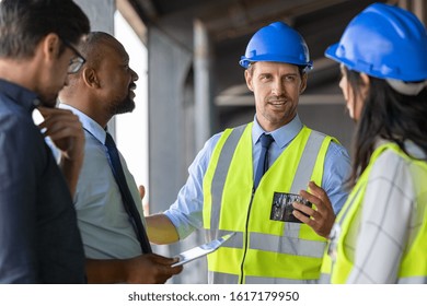 Happy Mature Engineer Discussing The Structure Of The Building With Architects Colleague At Construction Site. Engineers Wearing Safety Hardhat Having Work Conversation On The Safety Of The Structure.