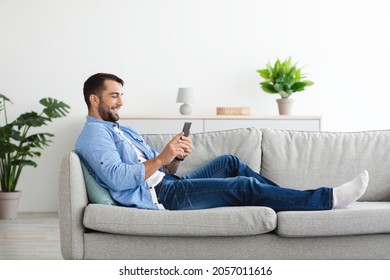 Happy Mature European Guy With Beard Sitting On Sofa And Typing On Phone, Play In Game In Living Room Interior, Copy Space. Free Time With App, Digital Gadget And Chatting Online In Social Networks