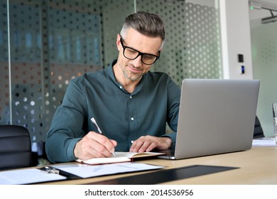 Happy mature executive ceo manager using laptop writing notes in notebook at workplace. Smiling middle aged business man working in office analyzing financial data doing research. - Powered by Shutterstock