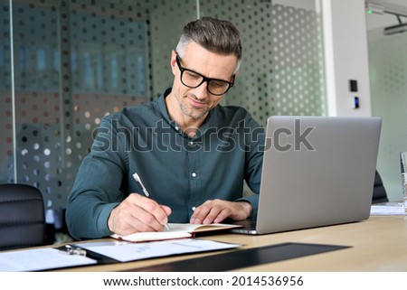 Happy mature executive ceo manager using laptop writing notes in notebook at workplace. Smiling middle aged business man working in office analyzing financial data doing research.