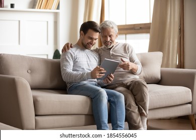 Happy Mature Father And Adult Son Using Tablet At Home Together, Smiling Older Grandfather And Grandson Looking At Device Screen, Sitting On Couch, Young Man Teaching Senior Dad To Use Computer