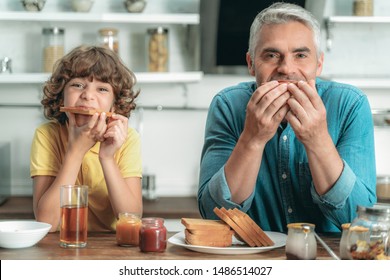 Happy Mature Father Sitting On Kitchen Near Little Son And Eating Toast Bread With Jam