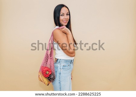Happy mature female in casual clothes carrying net bag with vegetables and fruits on shoulder and looking at camera with smile against beige background