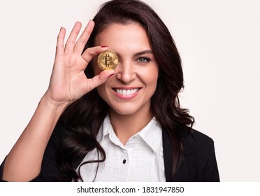 Happy mature female investor smiling for camera and holding bitcoin near eye against gray background