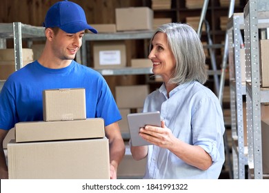 Happy Mature Female Manager Small Business Owner, Seller, Supervisor Using Digital Tablet Talking To Male Courier Holding Shipping Ecommerce Parcels Boxes Discuss Delivery Walking In Warehouse.