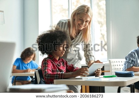 Happy mature female teacher educator helping African American junior school kid girl student using digital tablet computer education program app technology during elementary class lesson in classroom.