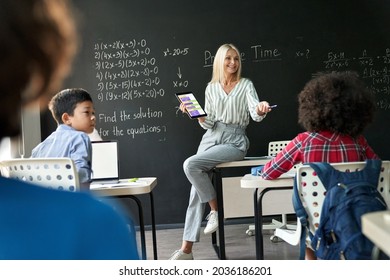 Happy Mature Female Teacher Teaching Multiethnic Kids At Maths Class In Junior Middle Elementary Education School Using Digital Tablet App Technology Talking To Students During Lesson In Classroom.