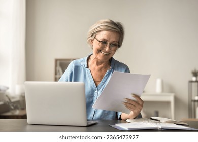 Happy mature freelance business woman in glasses reading good terms, conditions in legal document at home workplace, holding paper letter, smiling, laughing, sitting at laptop - Powered by Shutterstock