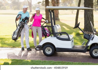 Happy Mature Golfer Couple By Golf Buggy While Walking On Field