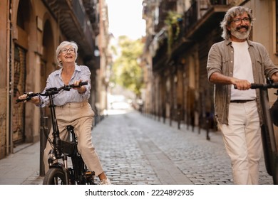 Happy Mature Gray Hair Couple In Push Scooter Riding Through The Old Town. Focus On Elderly Woman