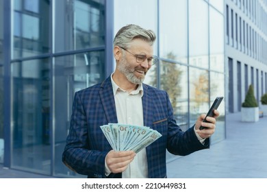 Happy Mature Gray-haired Businessman Holding Winnings Money Cash In Hands, Scared Man Smiling And Happy Looking At Smartphone Screen, Received Loan Outside Modern Office Building