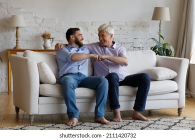 Happy Mature Grey Haired Dad Congratulating Adult Son On Success, Giving Friends Fist Bump Gesture, Support, Praise, Embracing Grownup Kid, Laughing, Sitting On Couch In Living Room