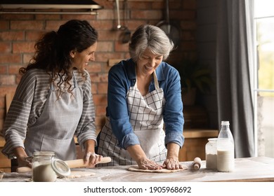 Happy mature grey haired woman with grownup daughter wearing aprons cooking handmade pastry, rolling dough, chatting, having fun, standing in kitchen at home, family spending leisure time together - Powered by Shutterstock