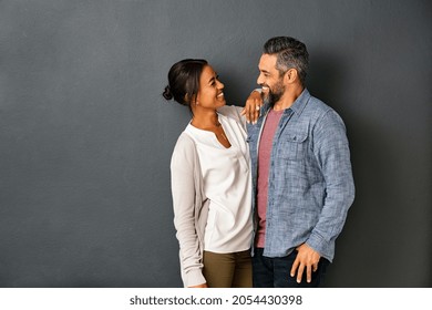 Happy Mature Indian Couple Embracing And Looking At Each Other Against Gray Background. Mid Adult Multiethnic Couple In Love Standing Against Grey Wall. Middle Eastern Man Fall In Love Of His Girl.
