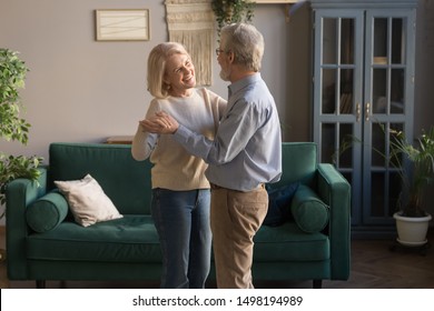 Happy Mature Loving Couple Dancing In Living Room Relaxing At Home On Weekend Together, Smiling Grey-haired Elderly Husband And Wife Spend Time Waltz Hugging Embracing, Spouses Enjoy Romantic Date