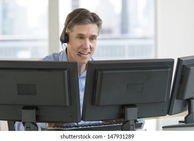 Happy mature male customer service representative using multiple screens at desk in office