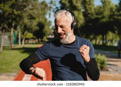 Happy mature male handsome runner listening to the music in headphones checking physical activity on fitness watch while jogging in stadium in the morning. Achieve goal. - Shutterstock ID 2182802943
