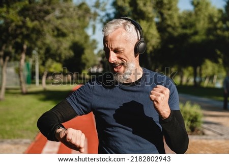 Happy mature male handsome runner listening to the music in headphones checking physical activity on fitness watch while jogging in stadium in the morning. Achieve goal. Foto stock © 