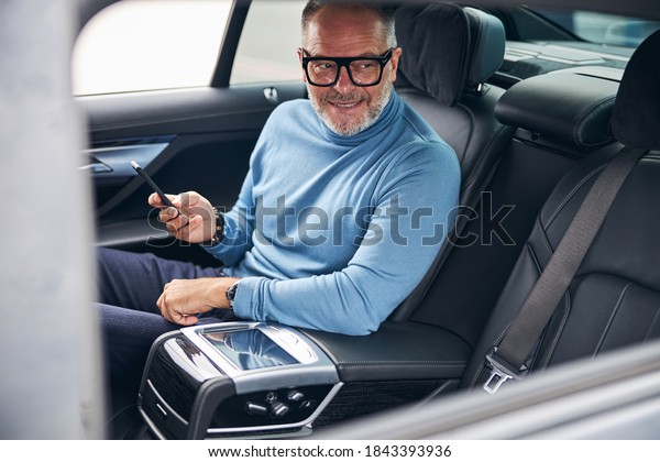 Happy mature male passenger with a cellular\
phone in his hand sitting in the\
taxi