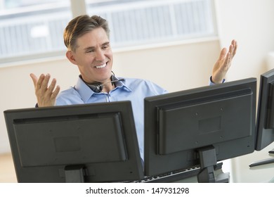 Happy Mature Male Trader Gesturing While Using Multiple Screens At Desk In Office