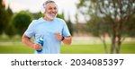 Happy mature man in headphones holding water bottle while jogging outside in park in early morning, senior sportsman running with beaming smile on his face enjoying active healthy lifestyle