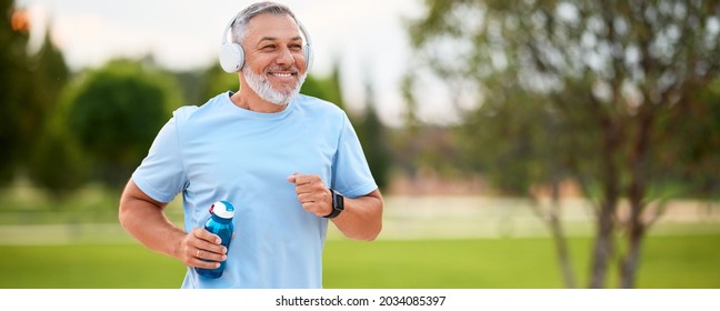 Happy mature man in headphones holding water bottle while jogging outside in park in early morning, senior sportsman running with beaming smile on his face enjoying active healthy lifestyle