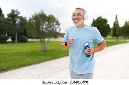 Happy Mature Man In Headphones Holding Water Bottle While Jogging Outside In Park In Early Morning, Senior Sportsman Running With Beaming Smile On His Face Enjoying Active Healthy Lifestyle
