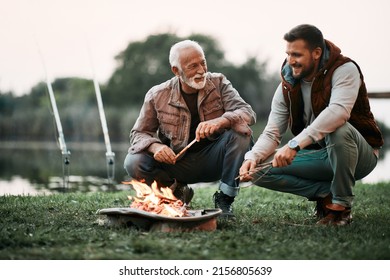 Happy mature man and his adult son talking while relaxing by campfire during their fishing day. - Shutterstock ID 2156805639