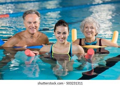 Happy Mature Man And Old Woman Doing Aqua Aerobics With Foam Rollers In Swimming Pool. Senior Couple Smiling With Swim Noodles Doing Aqua Fitness. Smiling Trainer With Mature Class Doing Aqua Gym.