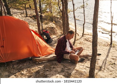 Happy mature man is sitting on cliff above sea and using notebook while having rest with tent