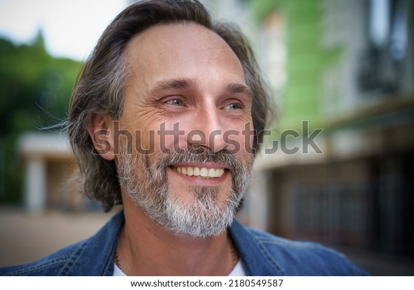 Happy mature man\
standing in city background smiling looking sideways. Handsome\
middle aged grey bearded man smiling on camera old town wearing\
casual. Travel concept. 