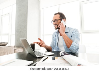 Happy Mature Man Talking On Mobile Phone While Sitting With Laptop Computer At The Table At Home