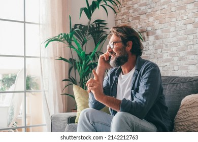 Happy mature man talking on mobile phone and gesturing while relaxing on couch in the living room of his house. Smiling caucasian guy using mobile phone sitting on sofa at home