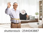 Happy mature man with a weight scale and a measuring tape gesturing thumbs up 