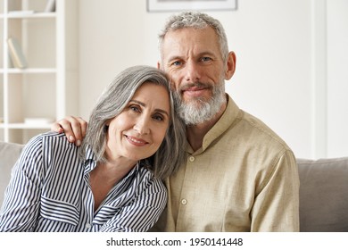 Happy Mature Mid Age Couple Hugging, Enjoying Bonding Sitting On Couch In Apartment. Smiling Affectionate Older 50s Husband Embracing Senior Wife Looking At Camera Relaxing At Home. Close Up Portrait