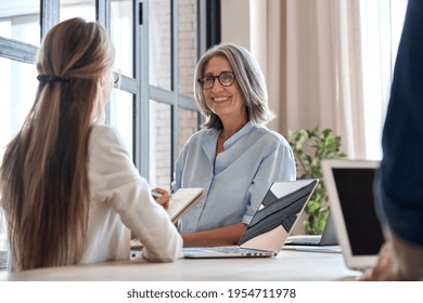 Happy Mature Middle 60s Aged Smiling Lady Boss Manager Teacher Talking To Assistant Or Student Meeting In Office Space. Company Team Working Together With The Laptops And Assistant Writing Notes.