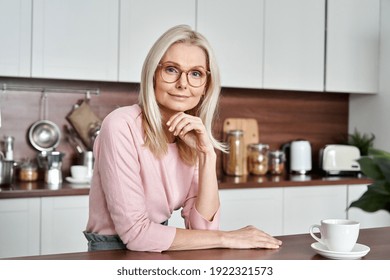 Happy Mature Middle Aged 50s Woman Wearing Glasses Sitting At The Kitchen Table. Smiling Older Adult Elegant Blond Lady Looking At Camera Posing At Home Drinking Coffee. Portrait.