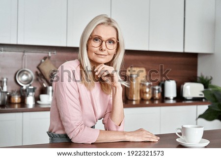 Happy mature middle aged 50s woman wearing glasses sitting at the kitchen table. Smiling older adult elegant blond lady looking at camera posing at home drinking coffee. Portrait. Stok fotoğraf © 