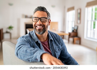 Happy mature middle eastern man wearing eyeglasses sitting on couch. Portrait of indian man relaxing at home and looking away with big smile. Mid adult guy with specs thinking about his future.