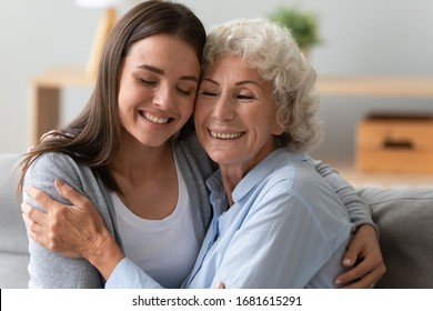 Happy Mature Mom And Grown-up Daughter Sit On Couch At Home Hug Cuddle Show Love And Care, Smiling Elderly Mother And Adult Girl Child Embrace In Living Room Enjoy Weekend Together