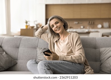 Happy mature naturally grey haired woman holding mobile phone, resting on sofa, looking at camera, posing indoors, smiling, laughing, using digital gadget for domestic Internet communication - Powered by Shutterstock